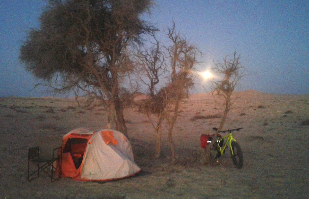 The sun rises in the background, and illuminates a lonely campsite with a single tent under an acacia tree.