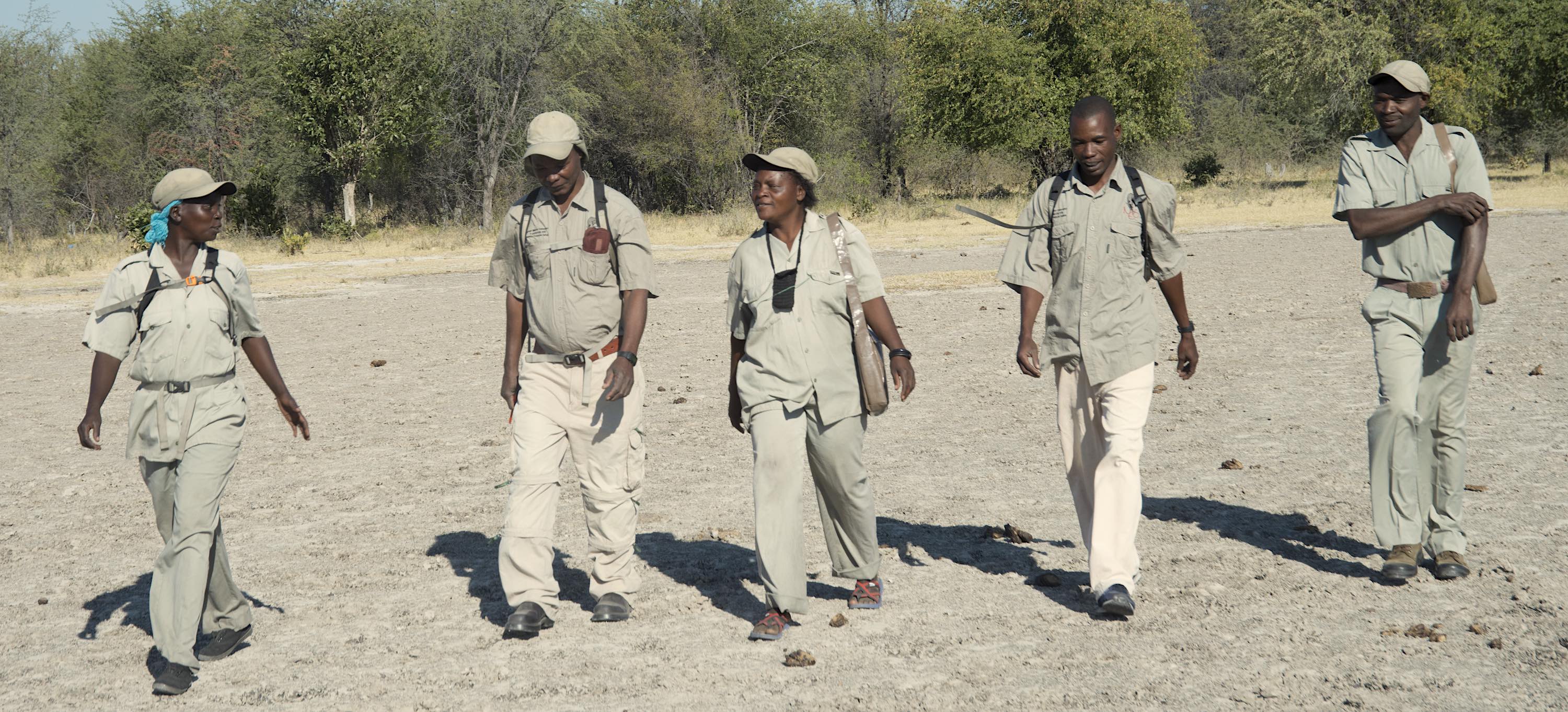 A group of five community game guards walk together across a dusty plain.