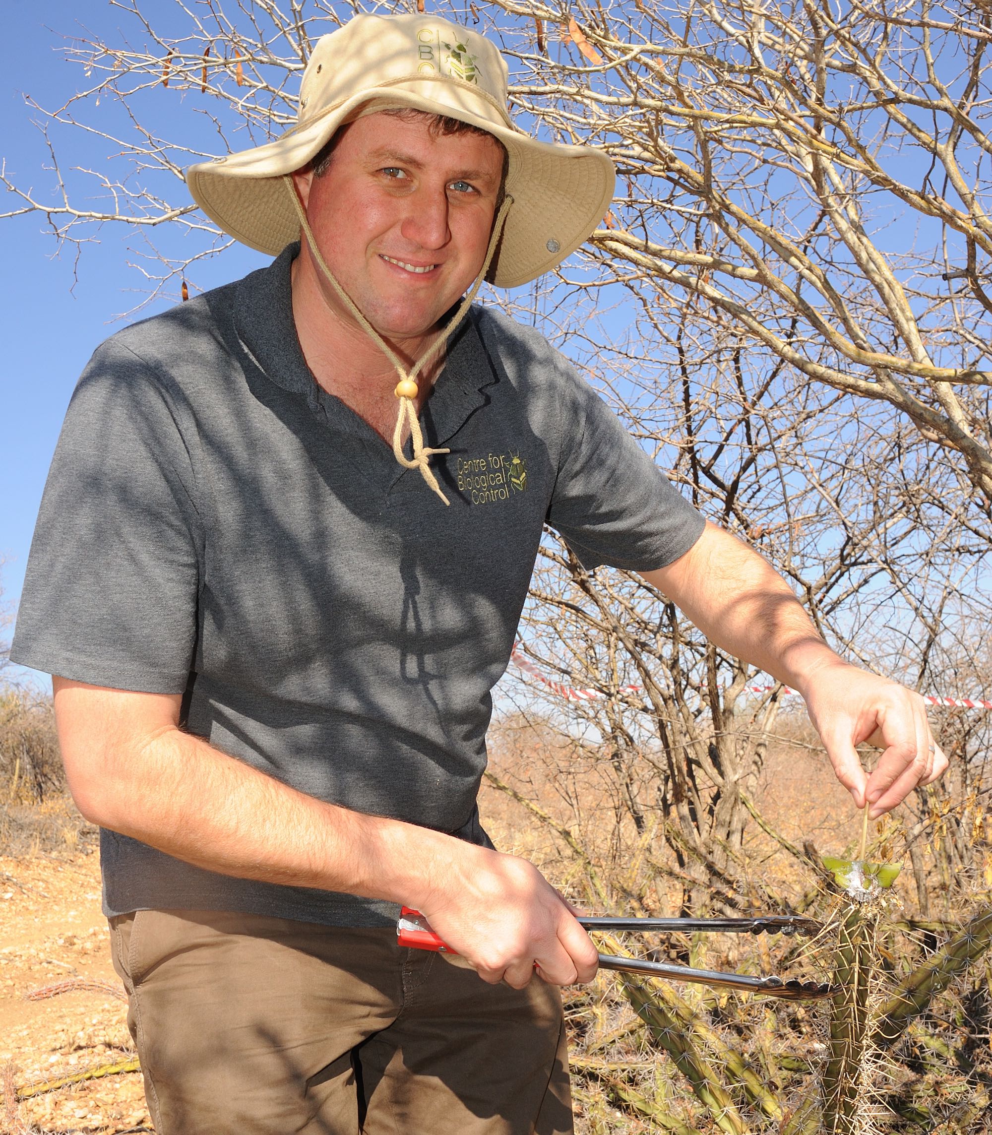 A man in a hat holding a cactus stem and a pair of braai tongs smiles at the camera.