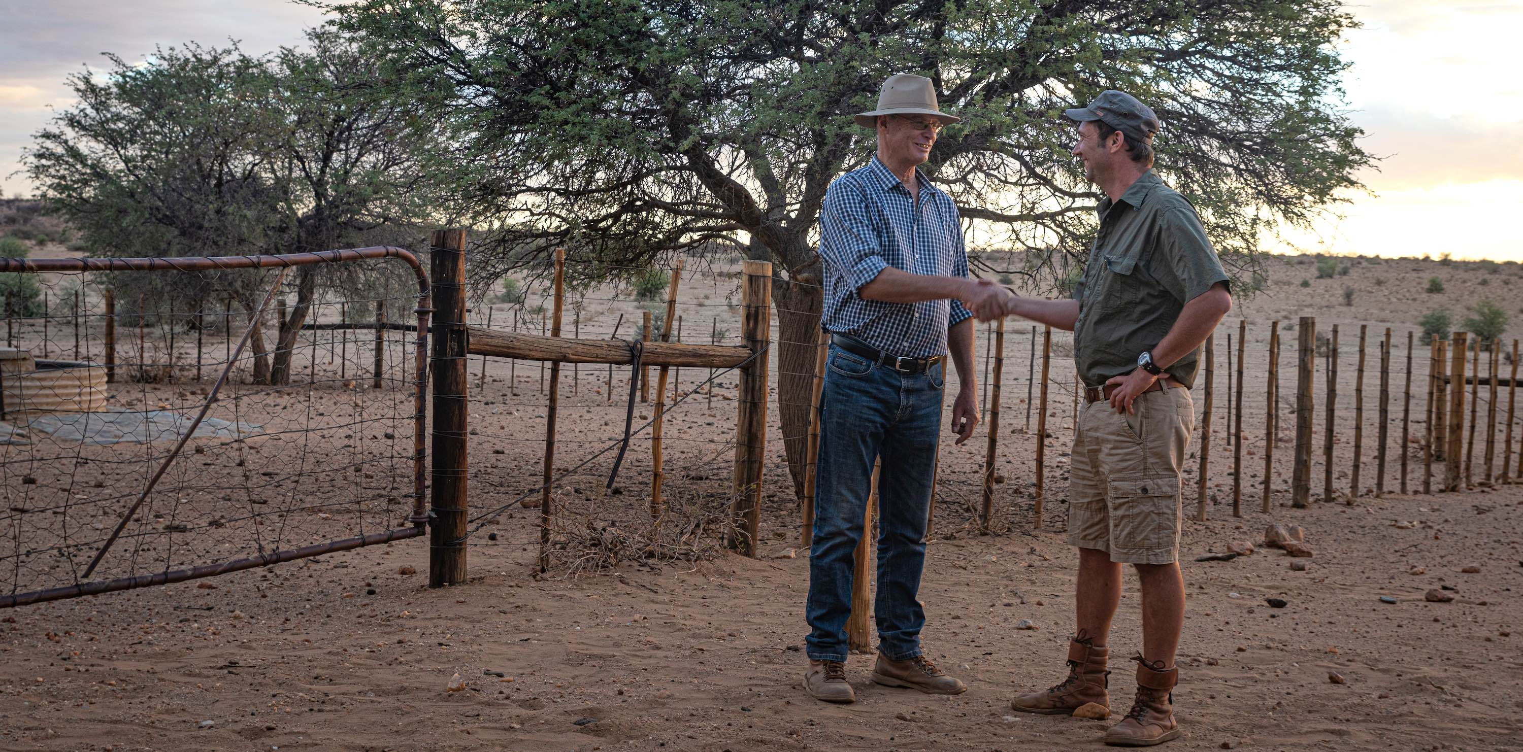 Two men shake hands next to a livestock fence.