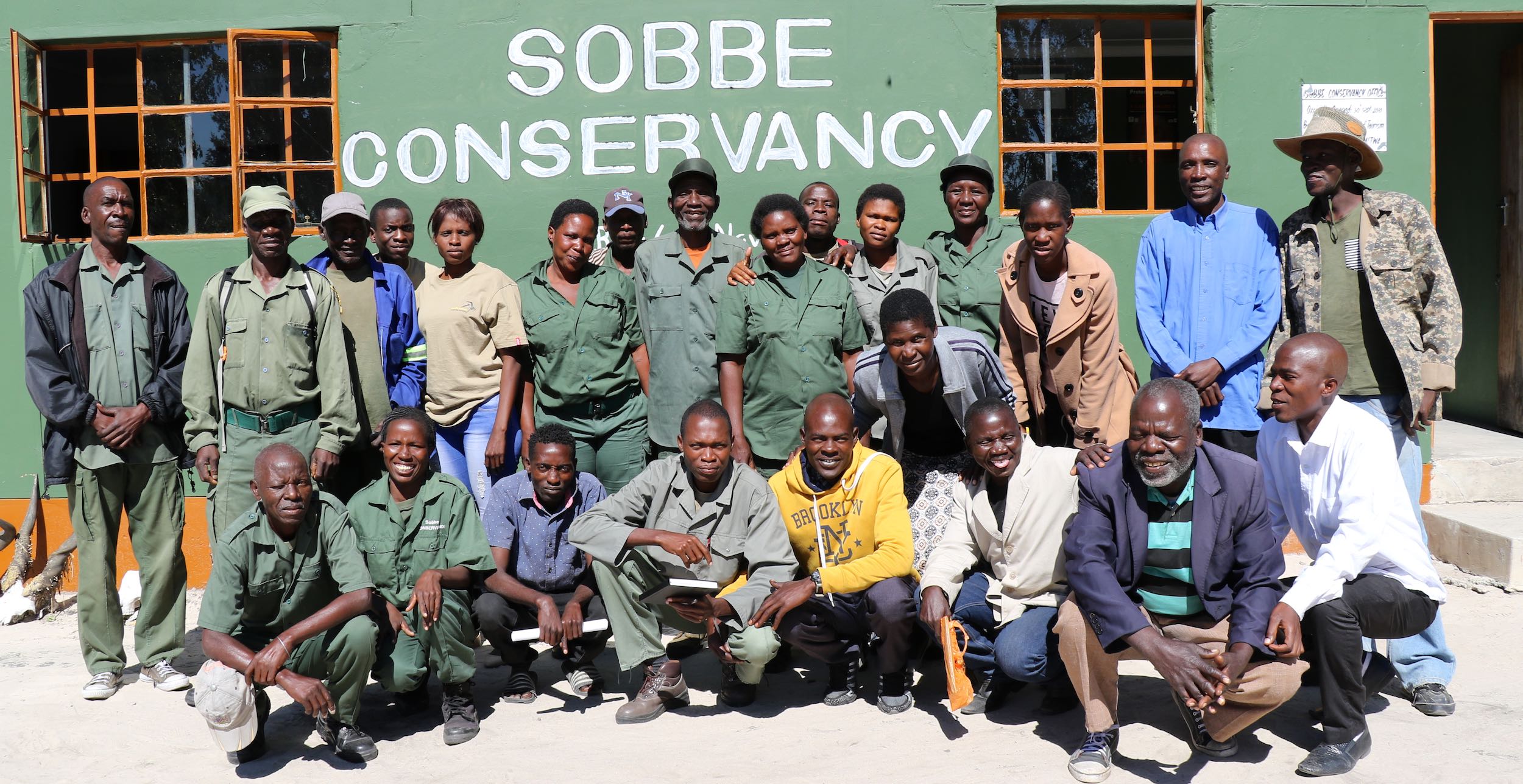 A group of Namibian men and women pose outside a green building.