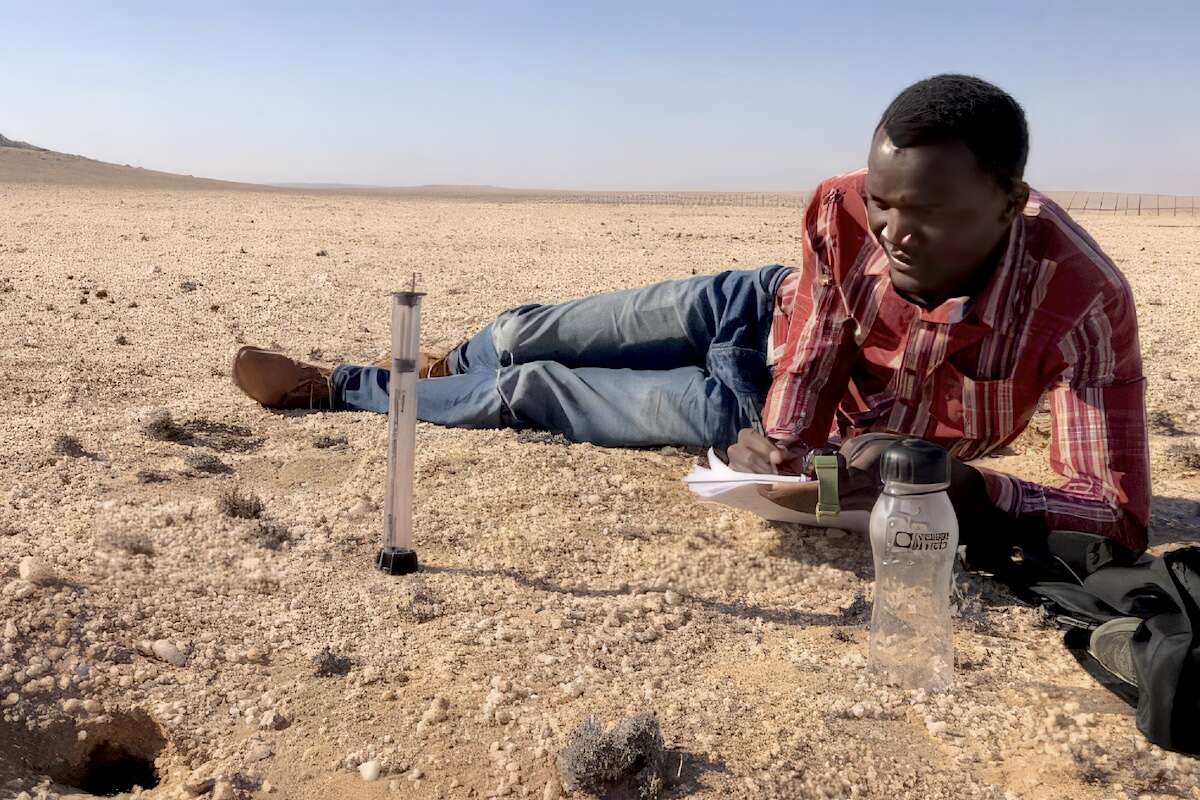 The author, with clipboard in hand sits next to an experiment site in the desert.