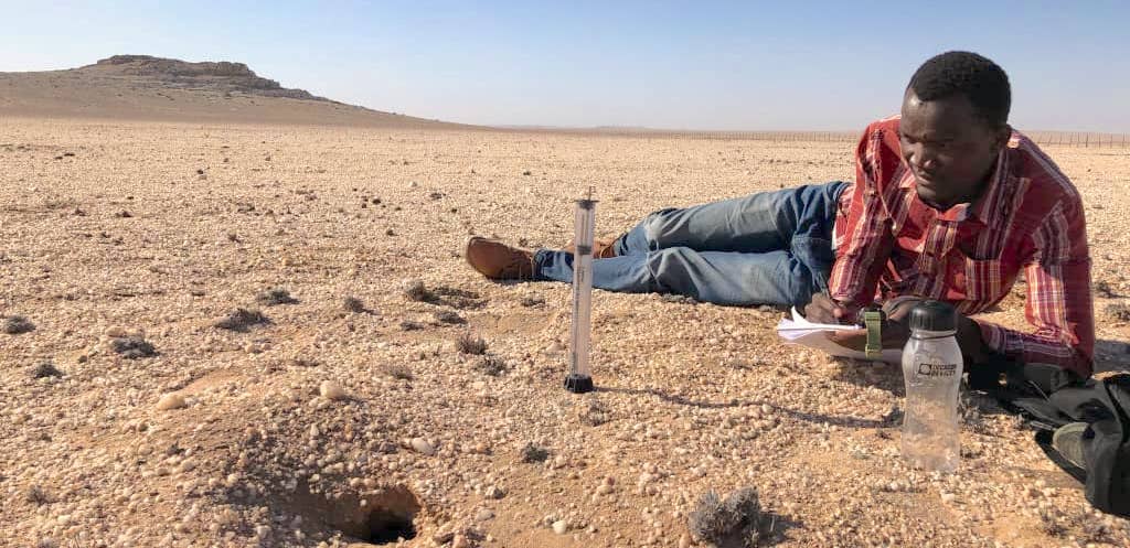 Halle Shaanika lies on his side in the desert recording information on a clipboard.