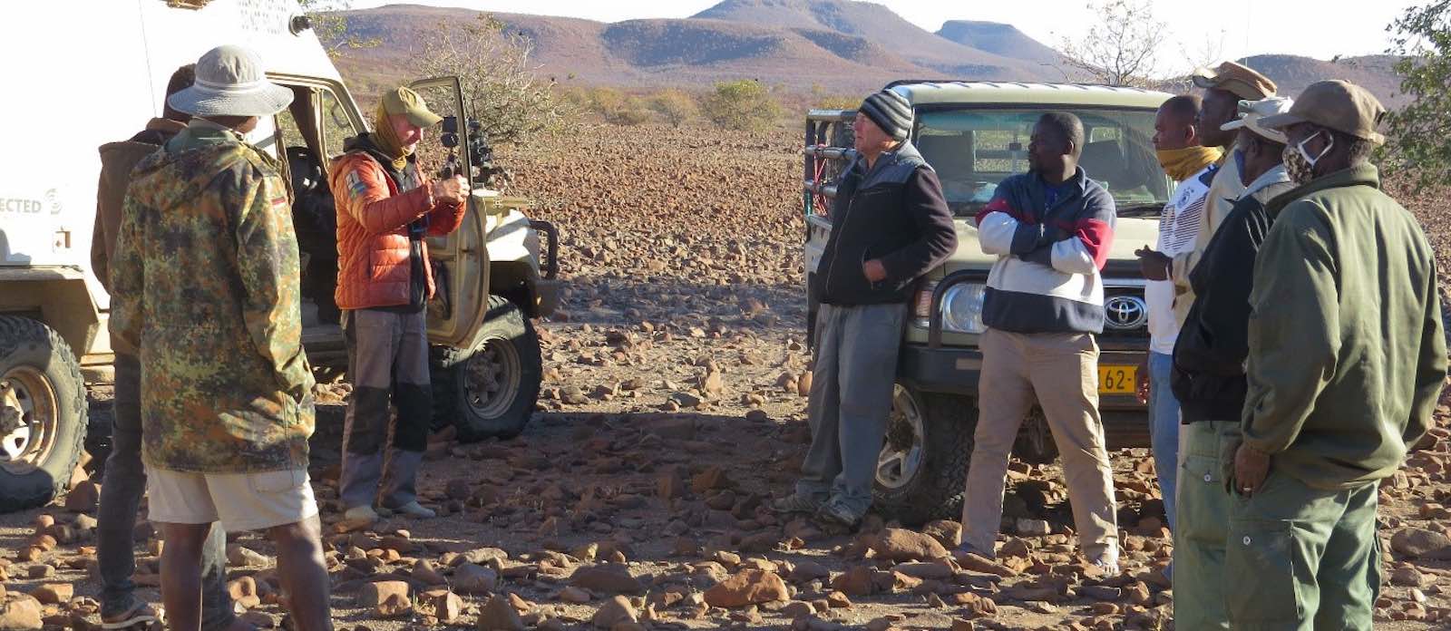 A group of people standing by two vehicles against a rocky and mountainous background.