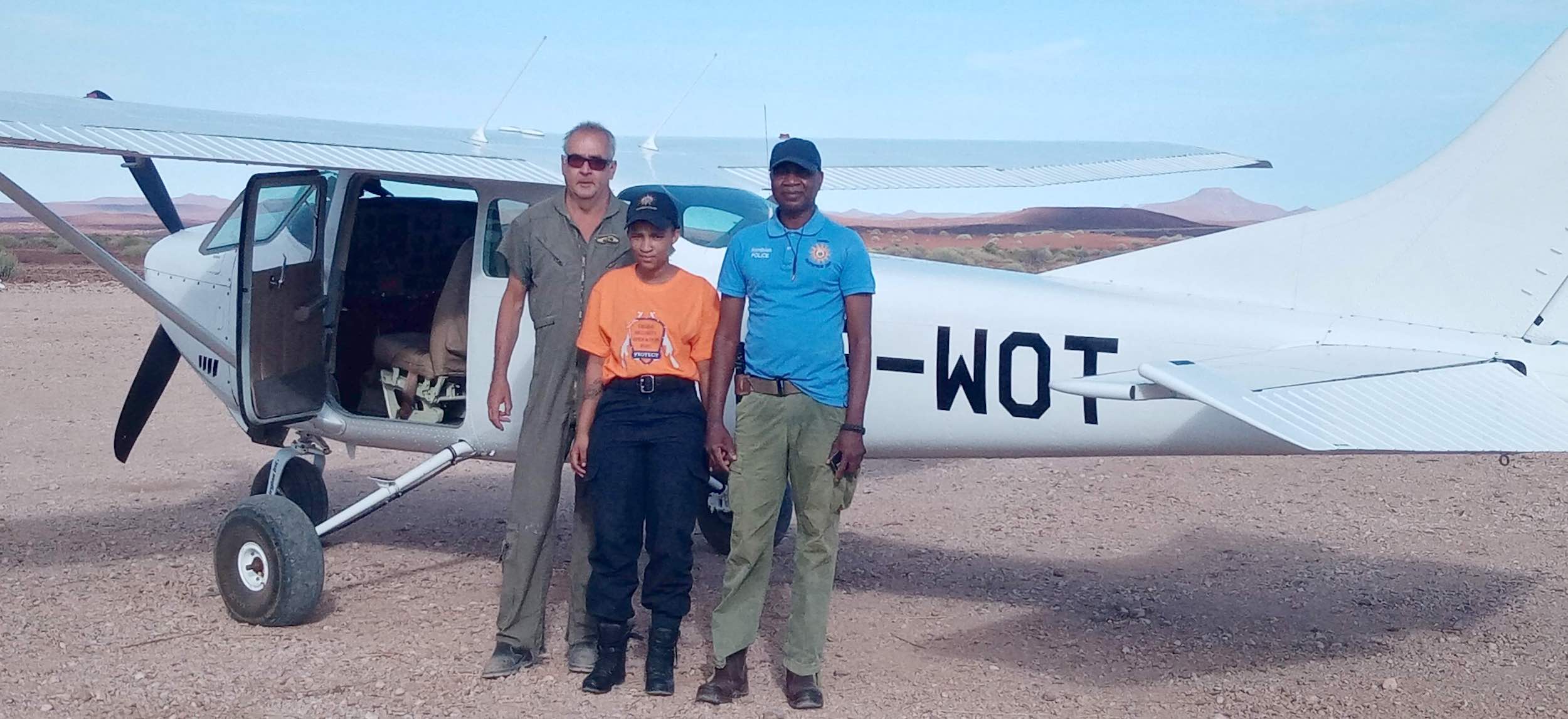 Two men and a woman stand in front of a Cessna light aircraft parked in the desert.