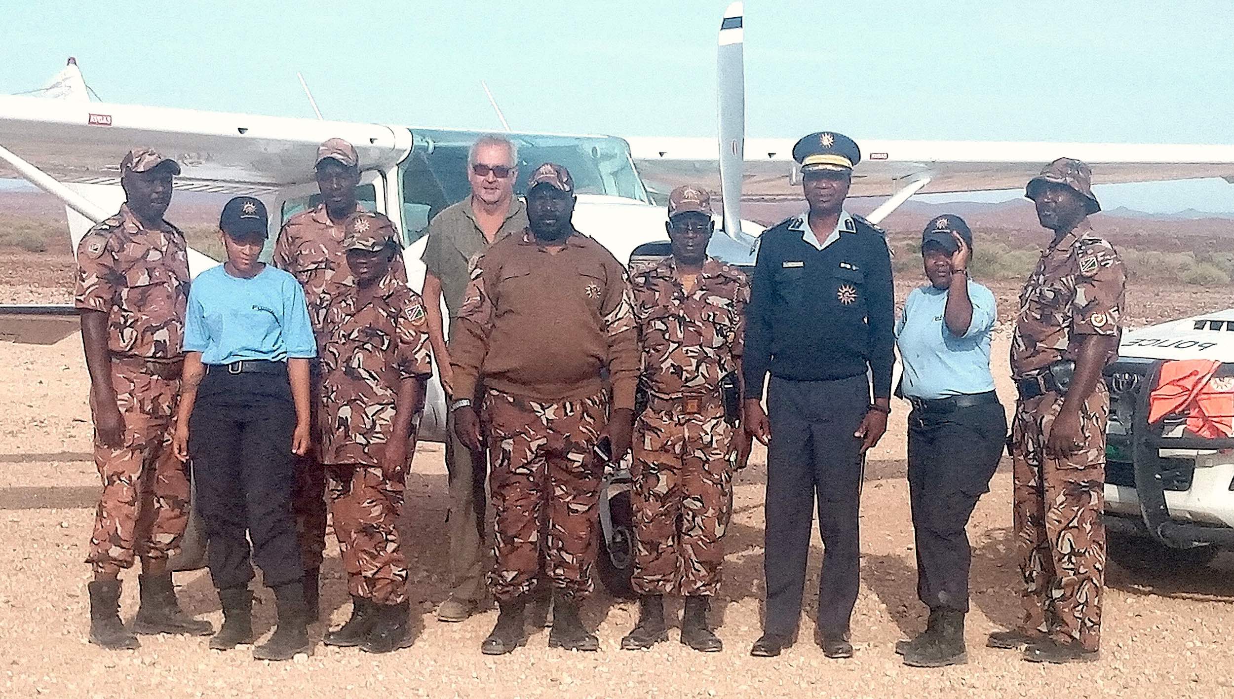 A group of eight people in uniform stand in front of a police vehicle and a light aircraft.
