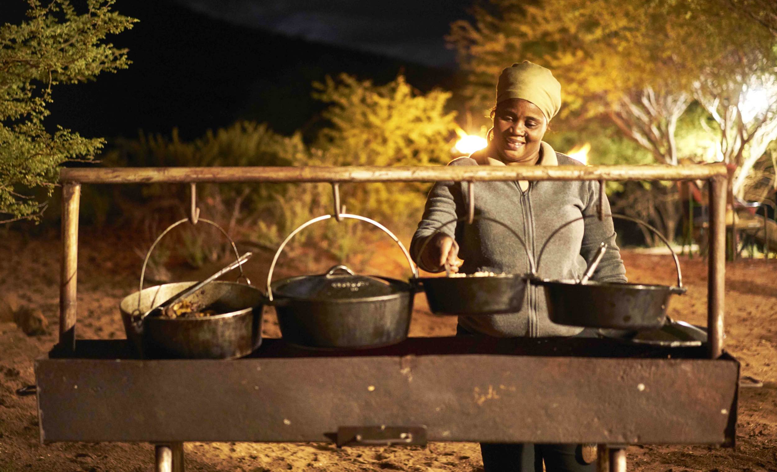 A woman prepares food in four iron pots hanging over an open fire.