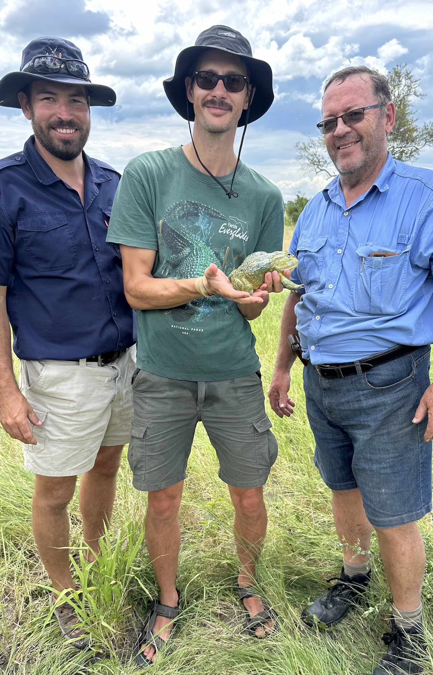 Three men smiling at the camera, while the middle one holds a bullfrog.
