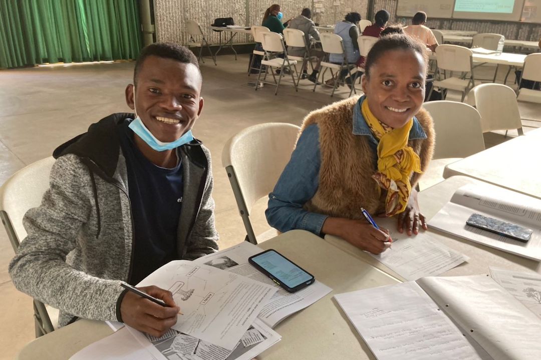 A young man and woman smile at the camera from behind the desk they are working upon.