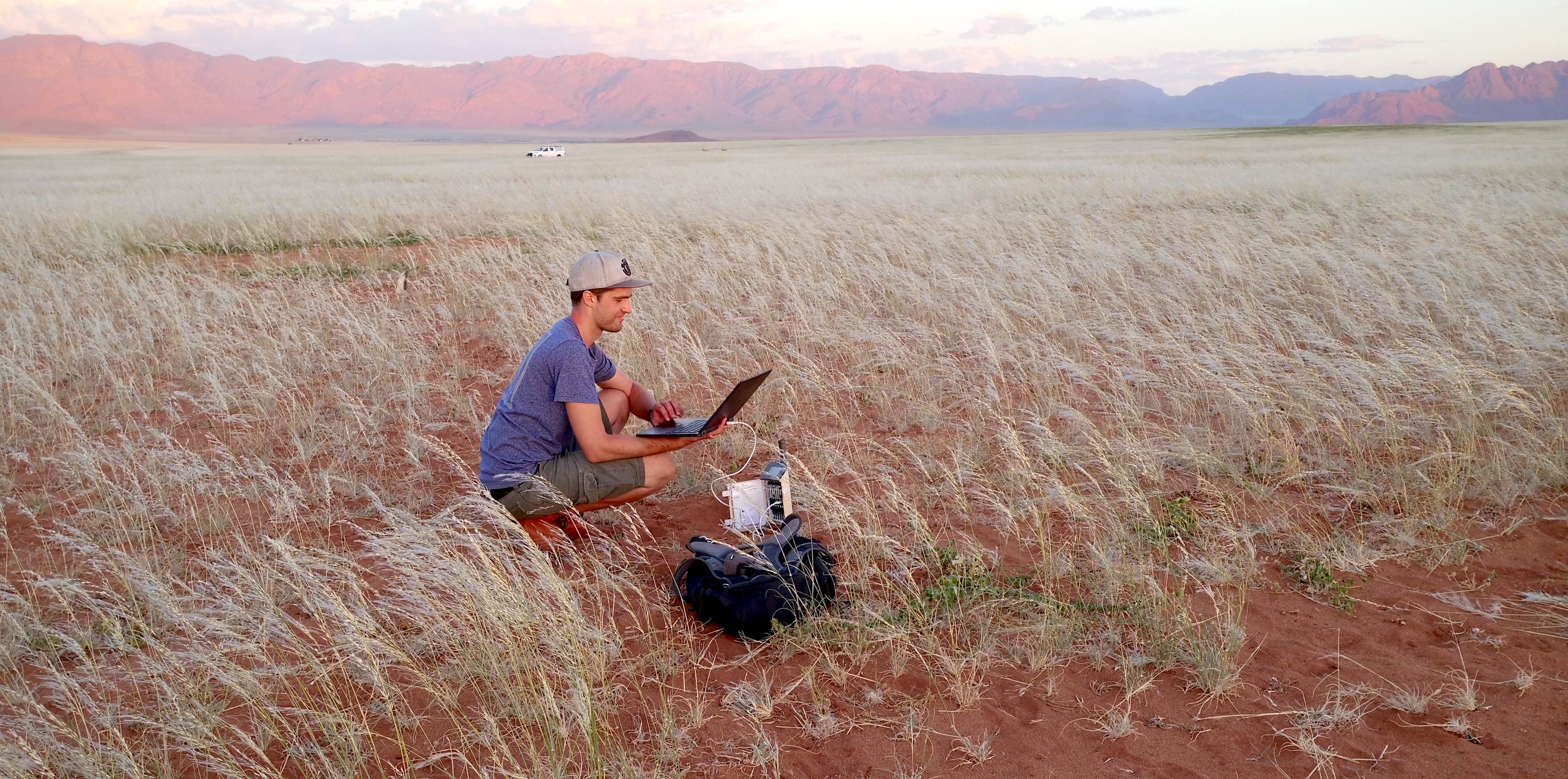 A man crouches in a field while holding a laptop.