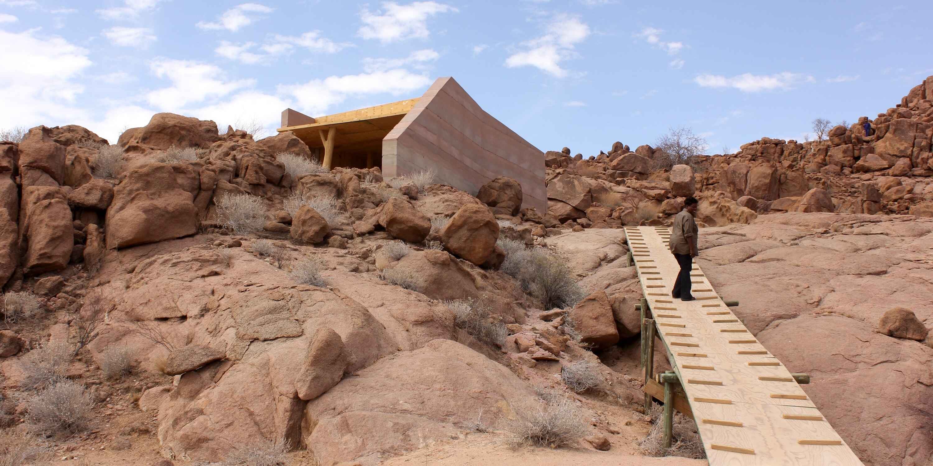 A smiling woman walks up a wooden path to a chalet among the rocks.