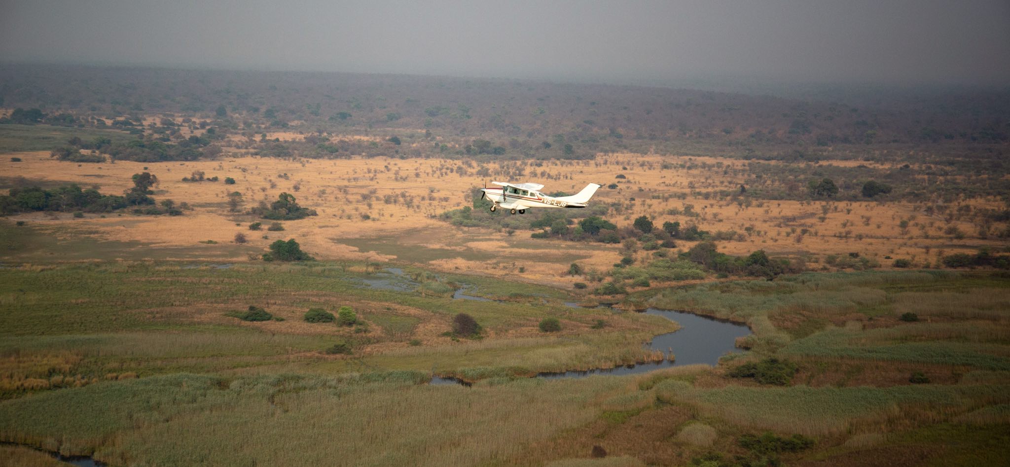 A Cessna light aircraft flying over a wide river in a green landscape.