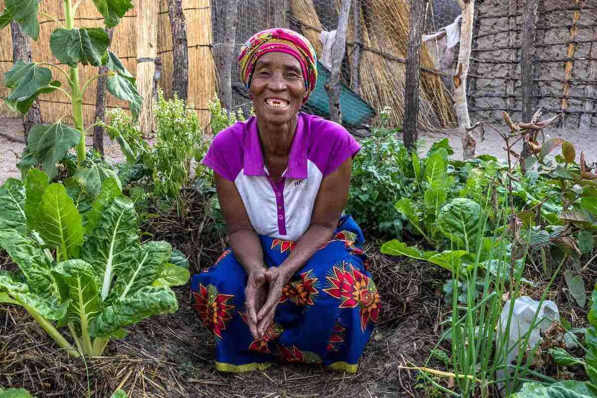 A woman sits smiling on the ground, surrounded by green plants.