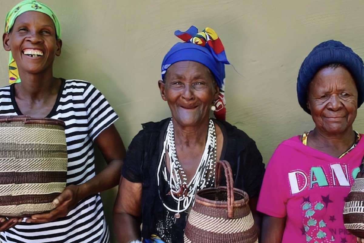 Three colourfully dressed San ladies holding woven baskets and smiling at the camera.