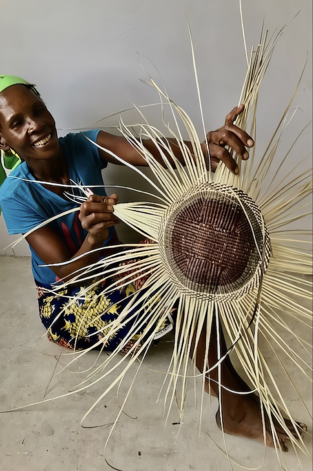 A San woman sits on the floor and shows a basket she is working on. It's at an early stage and looks much like a sunflower.