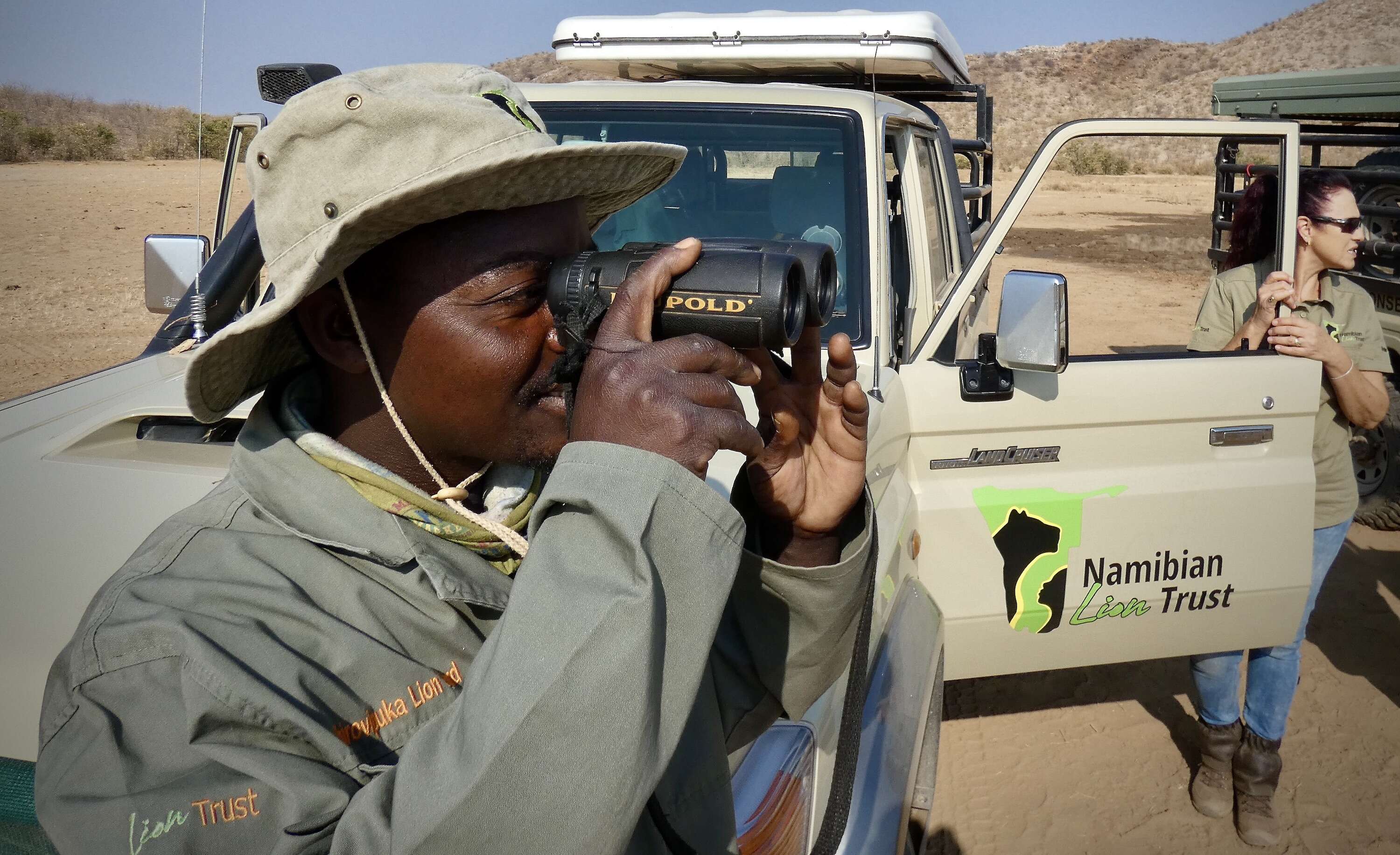 A lion ranger looks through binoculars while standing in front of an NLT bakkie.