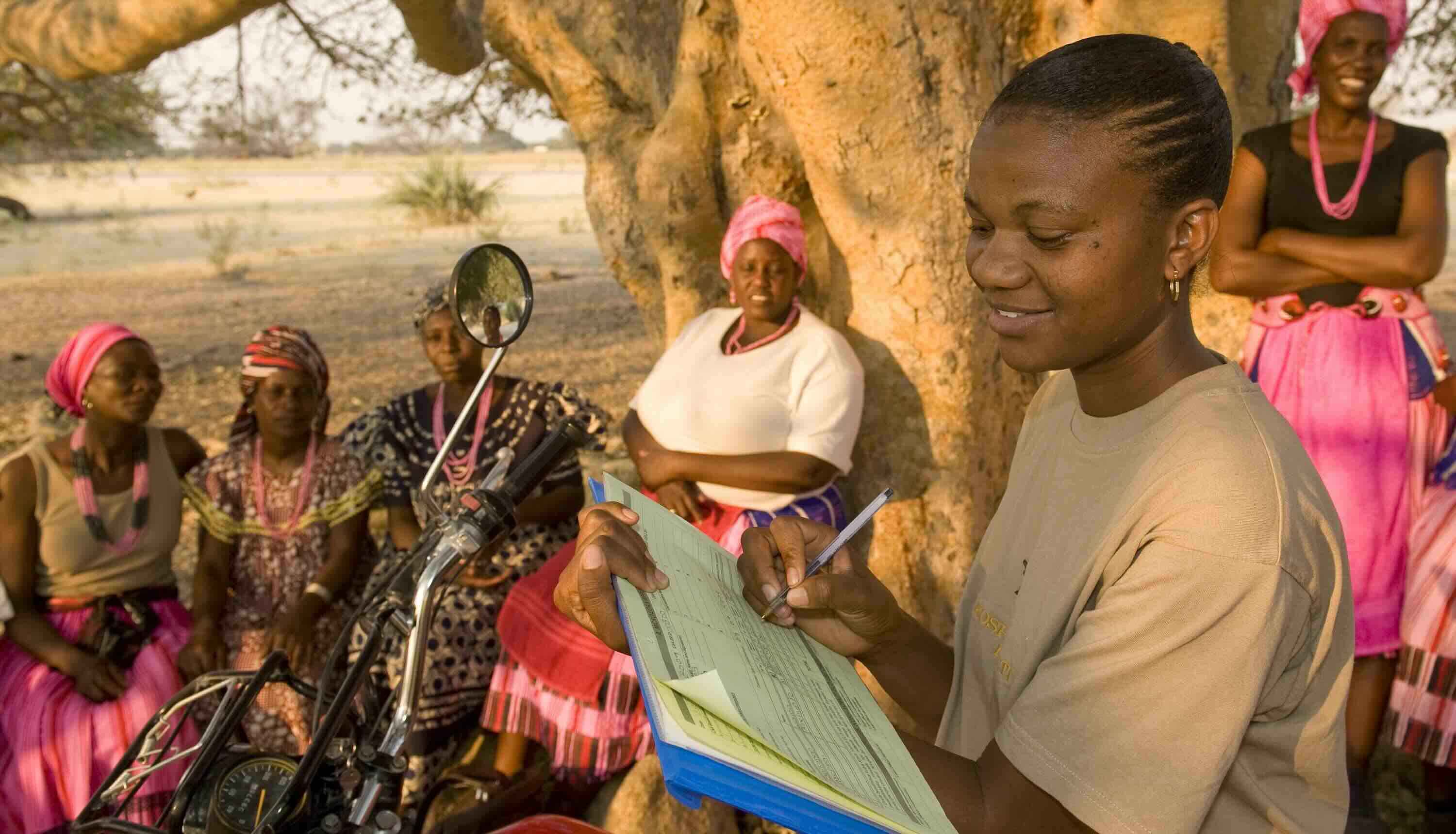 A woman on a motorcycle fills in data on a clipboard while several other woman sit under a tree beside her.