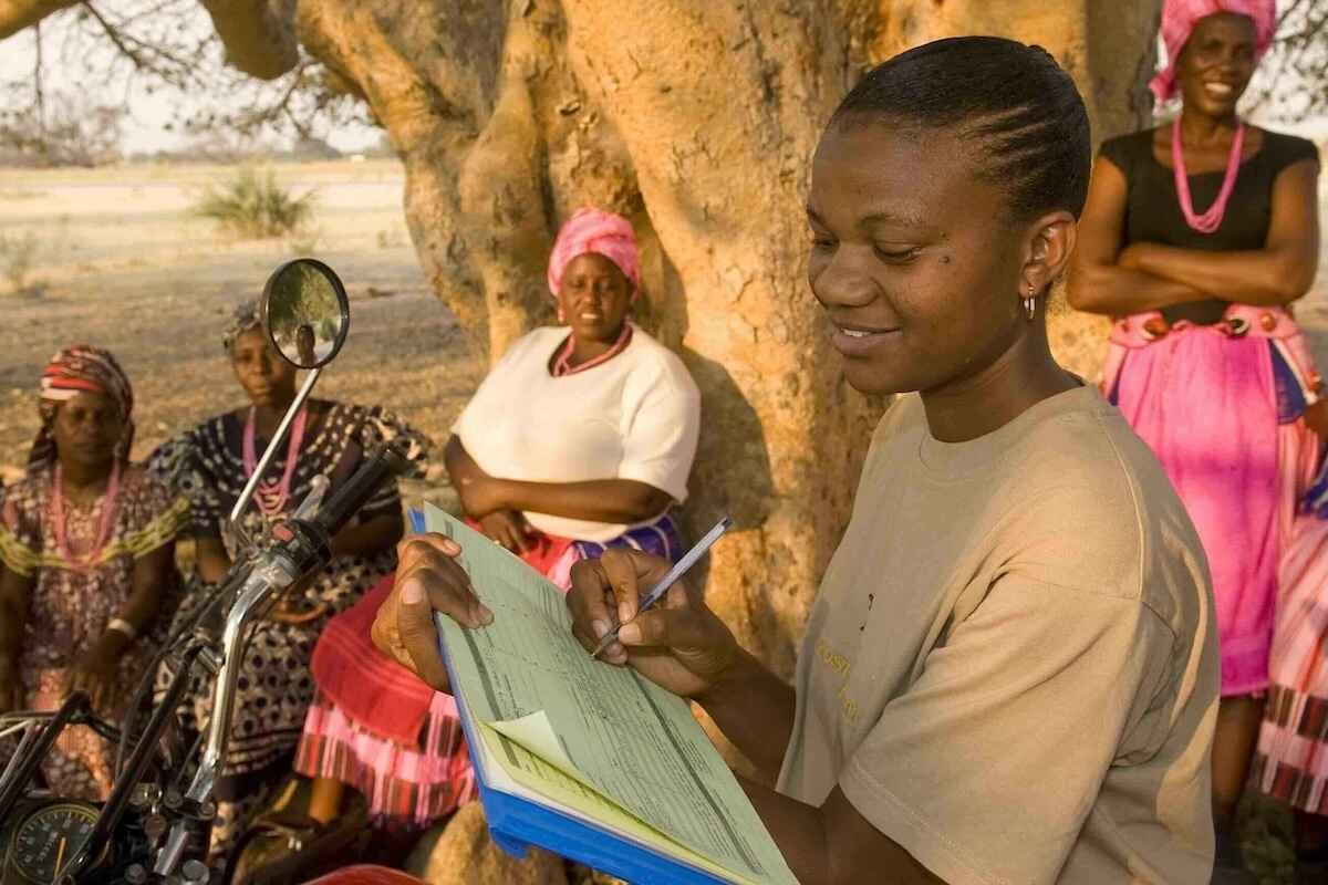 A woman on a motorcycle fills in data on a clipboard while several other woman sit under a tree beside her.