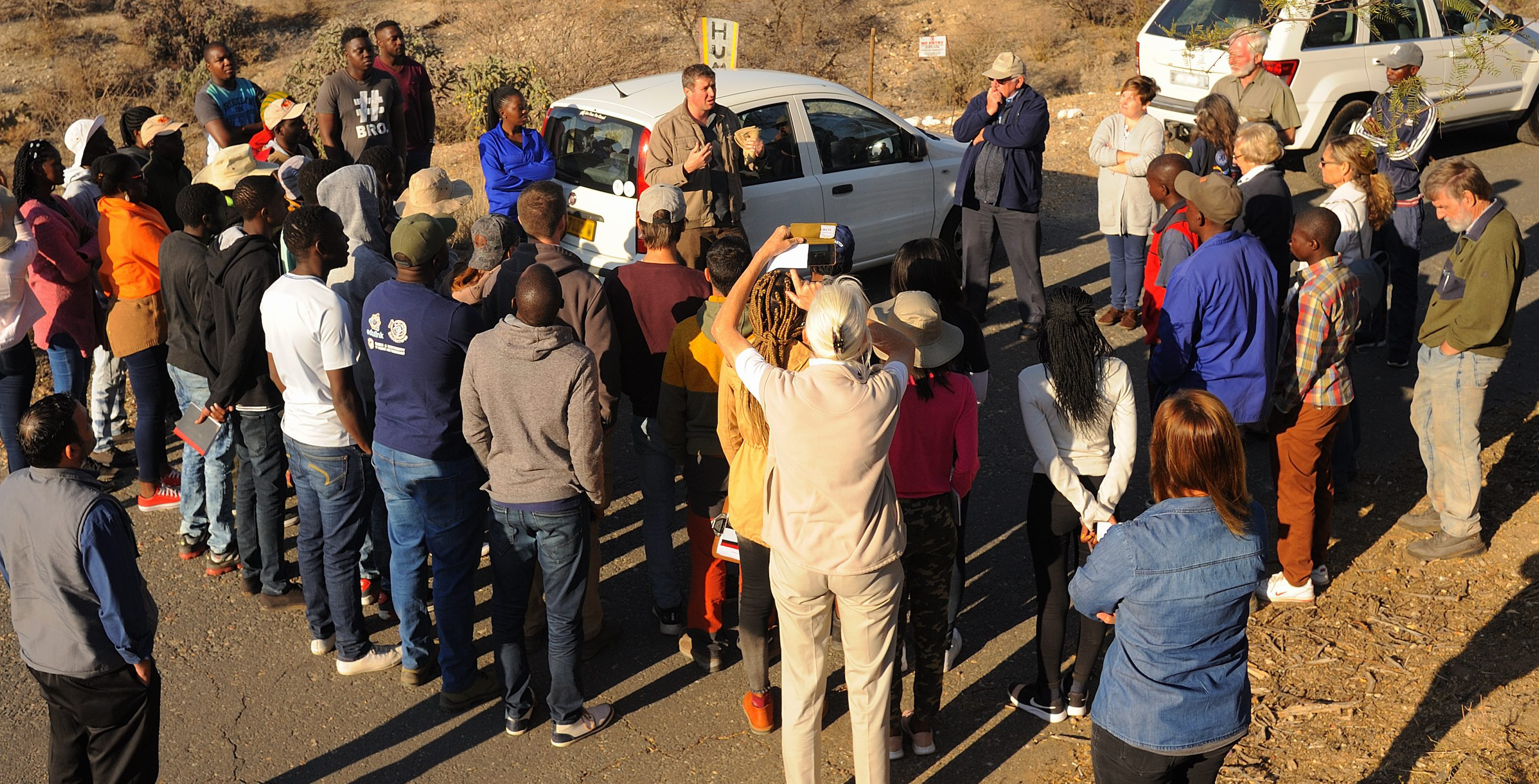 A group of people stand in a rough circle listening intently to a man talking to them.