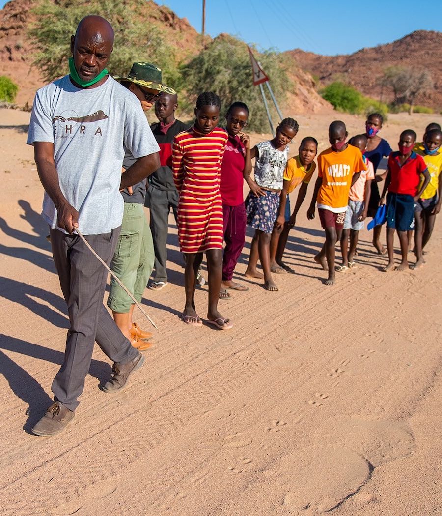 A man uses a stick to point towards various track on the sandy ground, while a group of children watch.