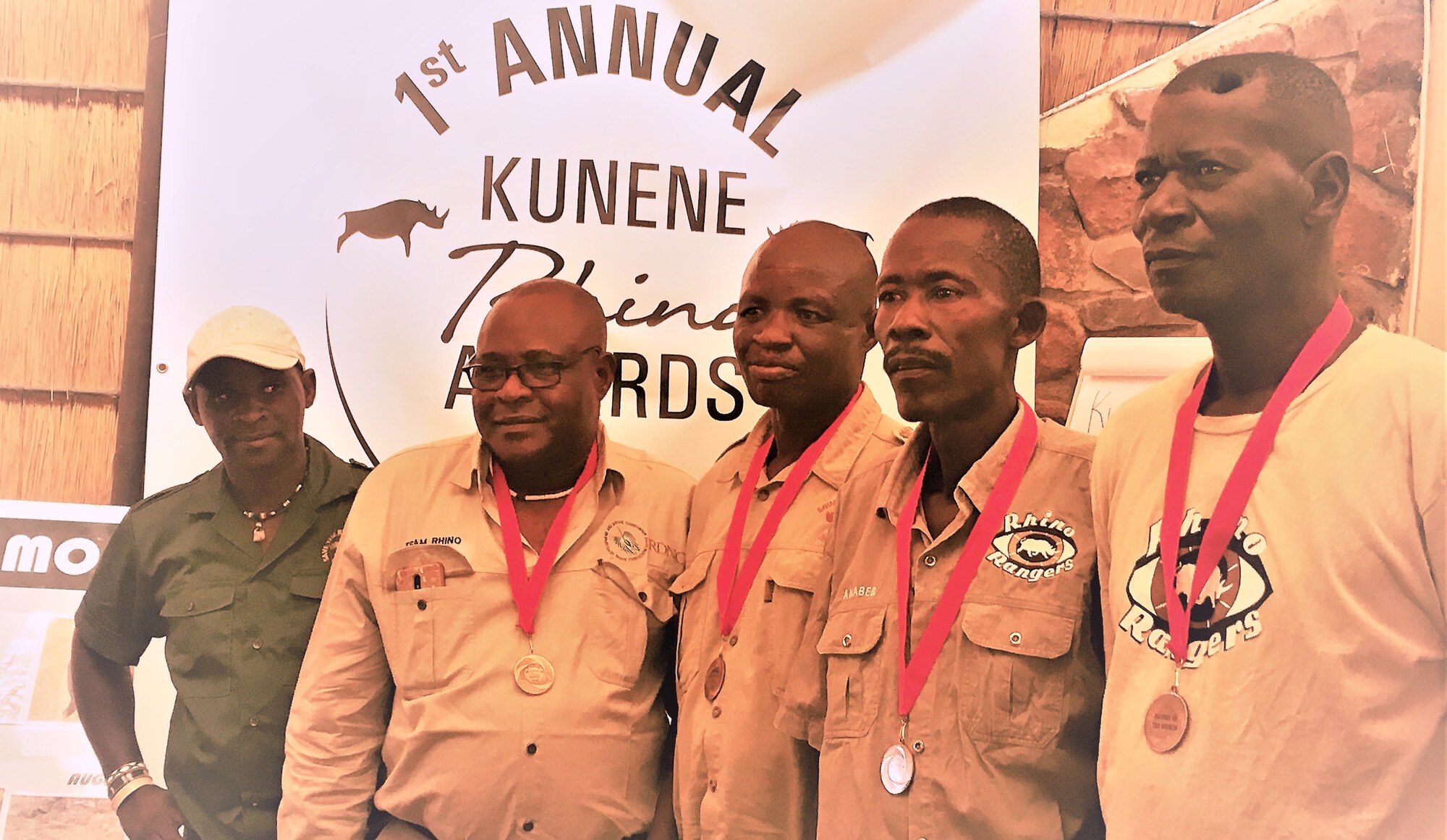 Four men stand side by side and wearing medals, while a fifth looks on.