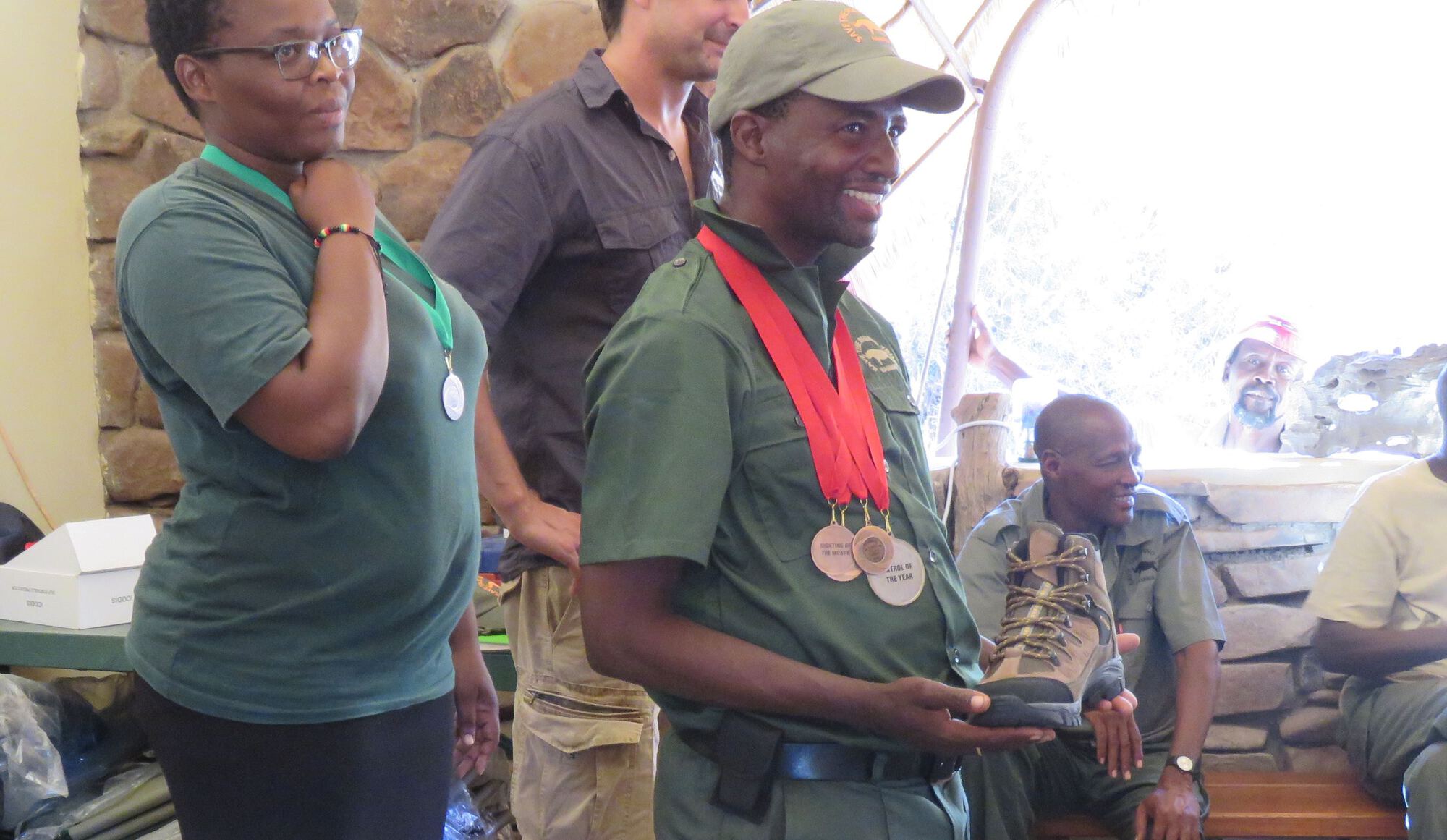 A man grins broadly as he holds his new boots, and proudly wears a collection of awards.