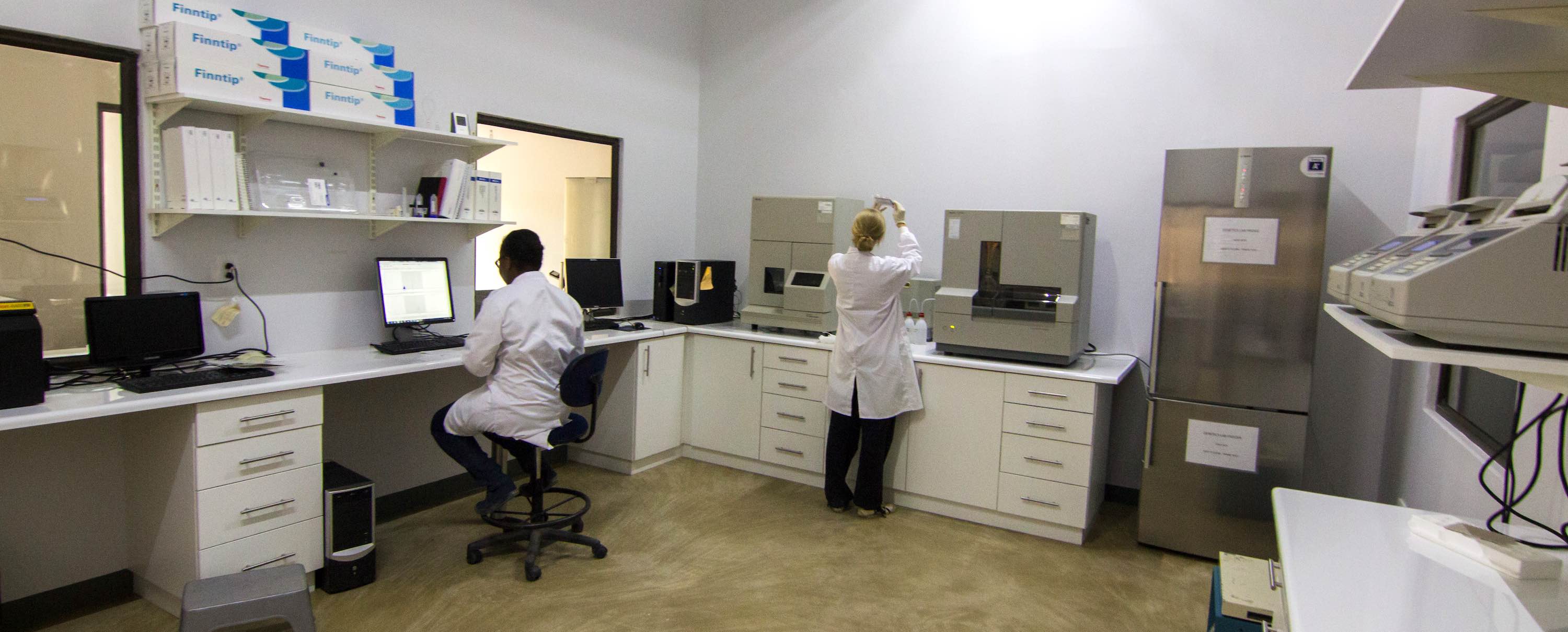 Two women with their backs to the camera working in a white-walled room dotted with various pieces of scientific equipment.