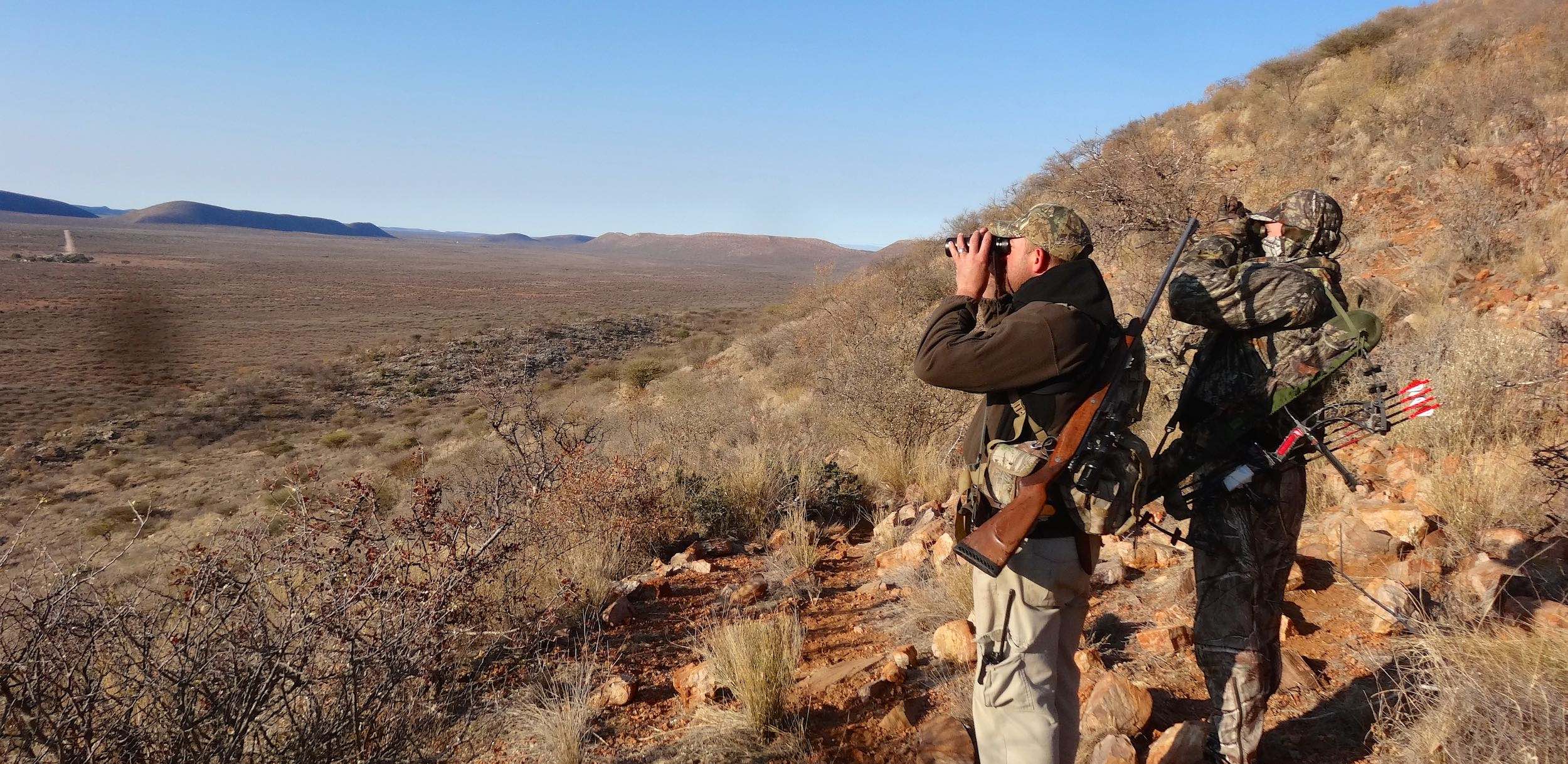 Two hunters on foot, one with a bow and one with a rifle, look for suitable prey.