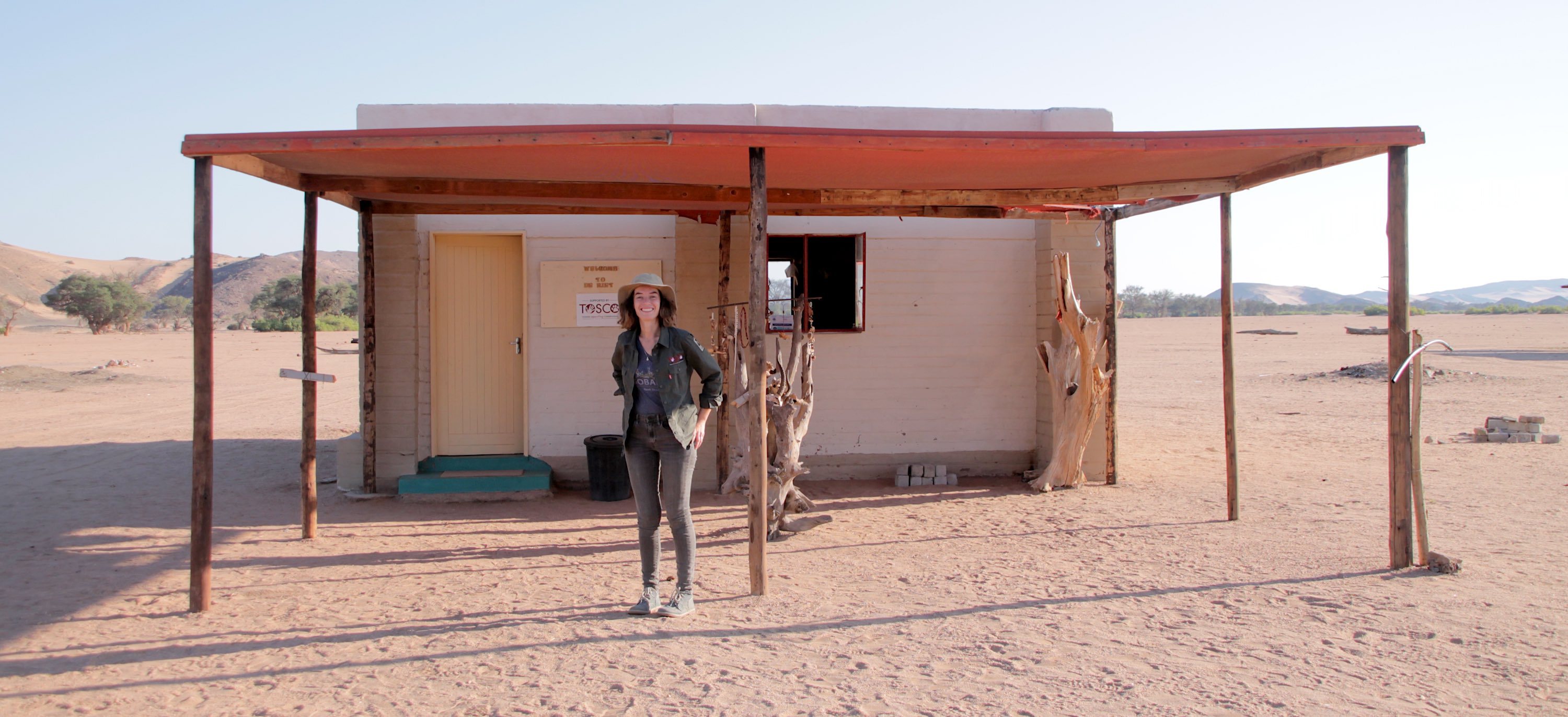 A woman stands in front of a simple office building in the midst of the desert.