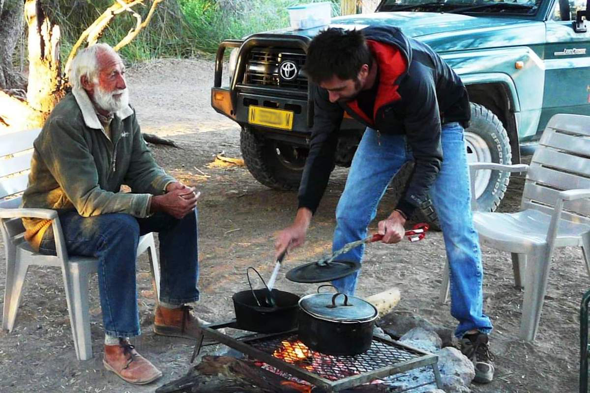 Garth Owen-Smith sits by a campfire while Felix Vallat cooks over it.