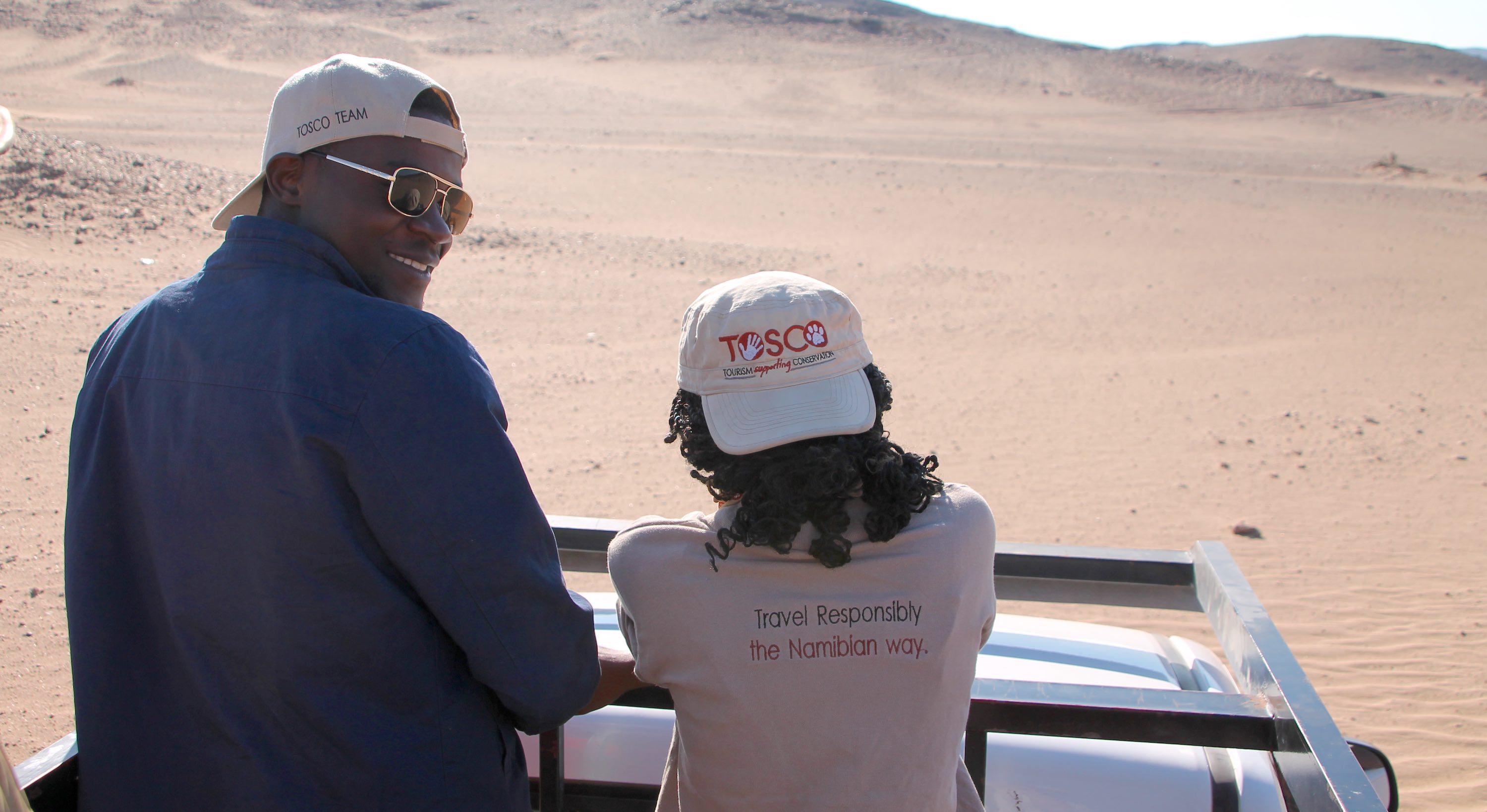 A man and a woman wearing TOSCO branded clothing riding in the back of a bakkie in the desert.